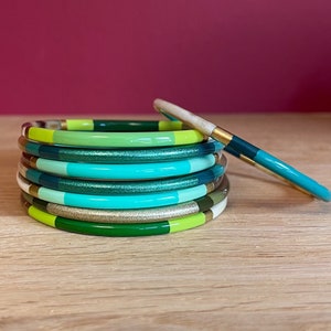 Puede incluir: Una pila de pulseras coloridas en tonos verdes, turquesa y dorado. Las pulseras están hechas de un material liso y brillante y tienen una forma ligeramente redondeada.