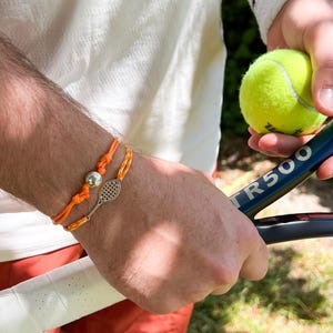Peut inclure: Deux bracelets orange avec des breloques argentées, l'un en forme de raquette de tennis et l'autre en forme de balle, sont portés au poignet. Une balle et une raquette de tennis sont tenues dans l'autre main. La raquette porte le texte "TR500".