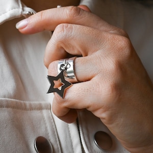 May include: A silver ring with a star-shaped charm. The ring is wide and has a smooth, polished finish. The star charm is open in the center and has a delicate chain connecting it to the ring.