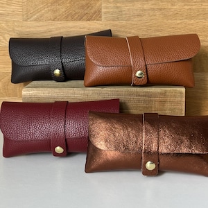 May include: Four leather eyeglass cases in different colours: dark brown, tan, burgundy, and metallic bronze. Each case has a flap closure with a strap and a gold-coloured snap. The cases are rectangular and made of textured leather.