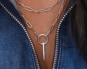 May include: A silver chain necklace with a long, thin pendant that has a circle at the top. The necklace is layered over a shorter silver chain.
