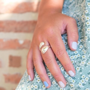May include: A silver-colored ring with a unique, flowing design. The ring features two rounded, teardrop-shaped elements that curve around the finger. The ring is worn on a hand with painted nails, and the background includes a floral-patterned dress.