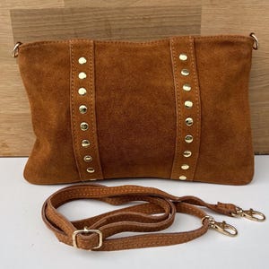 May include: A brown suede clutch with gold-colored rivets and a detachable shoulder strap. The bag has a rectangular shape and a soft texture. The strap is also brown and has gold-colored clasps.