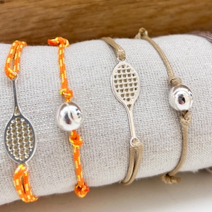 Puede incluir: Una colección de pulseras con temática de tenis. Las pulseras presentan dijes plateados con forma de raquetas y pelotas de tenis. Los cordones son de varios colores, incluyendo naranja, beige y gris. Las pulseras se exhiben sobre una tela gris claro.
