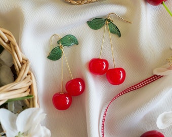 May include: Gold cherry earrings with green leaves. The earrings are on a white fabric background with red and white checkered ribbon and red cherries.