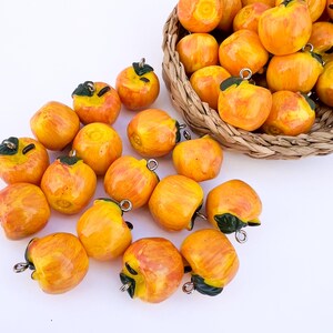 Peut inclure: Une collection de petites breloques rondes en forme de pomme orange avec des feuilles vertes et des anneaux en métal argenté pour les suspendre. Certaines breloques sont disposées dans un panier en osier, d'autres sur une surface blanche.