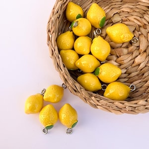 Peut inclure: Des breloques en forme de citrons jaunes avec des détails de feuilles vertes et des boucles argentées. Certains sont dans un panier en osier, d'autres éparpillés à l'extérieur. Les citrons ont une surface texturée.
