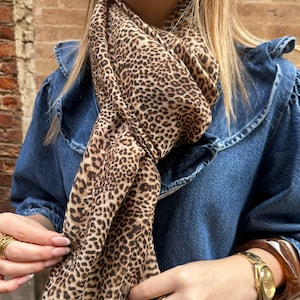 Peut inclure: Écharpe à imprimé léopard marron et beige autour du cou. Le foulard est fait d'un tissu léger et présente un motif animalier classique. La personne porte une veste en jean.
