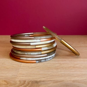 Könnte beinhalten: Ein Stapel von Armreifen in einem Farbkonzept aus Weiß, Gold und Kupfer. Die Armreifen sind aus einem glatten, glänzenden Material gefertigt und haben ein schlichtes, elegantes Design.