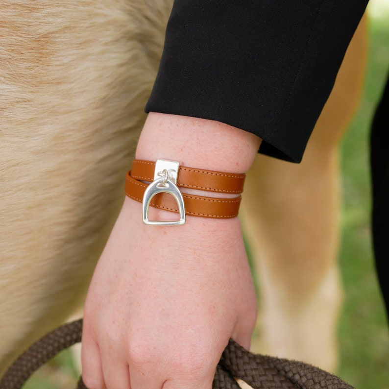 Pode incluir: Um bracelete de couro marrom de dupla volta com um fecho de estribo prateado. O bracelete &eacute; usado no pulso.