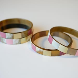 Könnte beinhalten: Drei übereinanderliegende Armreifen mit einem geometrischen Muster aus rosa, goldenen und weißen Quadraten.