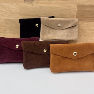 Può includere: Una collezione di pochette in camoscio in vari colori: nero, beige, bordeaux, marrone e ruggine. Ogni pochette ha una chiusura a patta assicurata da un bottone a pressione dorato. Le pochette sono disposte su una superficie chiara.