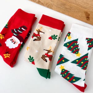 Op de afbeelding: Drie paar kerstsokken. Eén paar is rood met een kerstmanontwerp, een ander is beige met rendieren en het derde is wit met kerstbomen. Elk paar heeft een contrasterende boord en teen.