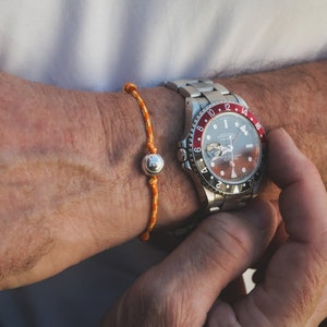Puede incluir: Un reloj plateado con una esfera negra y detalles rojos. El reloj tiene un bisel giratorio con los números del 1 al 24. El reloj está en la muñeca de una persona con una pulsera naranja y blanca.