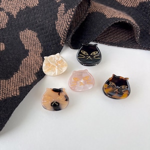 Puede incluir: Cinco pinzas para el pelo con forma de gato en varios colores, incluyendo negro, carey, rosa y crema. Cada pinza presenta un diseño detallado de cara de gato con detalles dorados o negros. Una bufanda estampada está en el fondo.