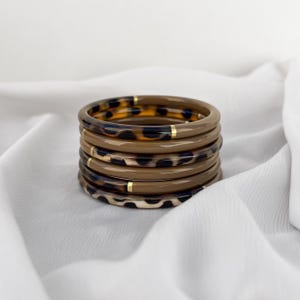 Può includere: Una pila di sei braccialetti marroni con motivi a tinta unita e leopardati. Ogni braccialetto ha un accento dorato. I gioielli sono esposti su un tessuto bianco strutturato.