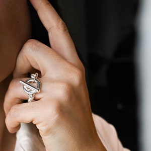 Puede incluir: Un anillo de plata con un diseño de cierre de palanca. El anillo se lleva en el dedo índice de una mano.