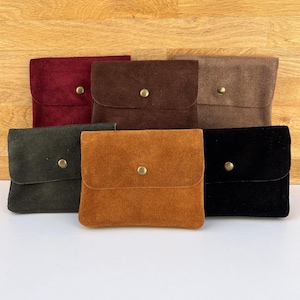 May include: A collection of six small, suede pouches in various colors: burgundy, brown, tan, olive green, orange, and black. Each pouch has a flap closure secured by a gold-colored snap. The pouches are arranged on a light-colored surface.