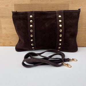 May include: A dark brown suede handbag with gold-colored stud accents and a matching shoulder strap. The bag has a rectangular shape and a zipper closure. The strap is detachable with gold-colored clasps.