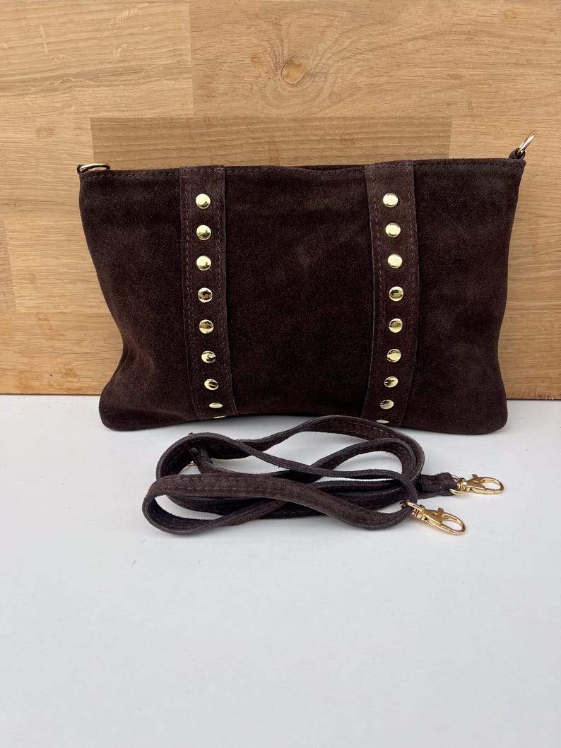 May include: A dark brown suede handbag with gold-colored stud accents and a matching shoulder strap. The bag has a rectangular shape and a zipper closure. The strap is detachable with gold-colored clasps.