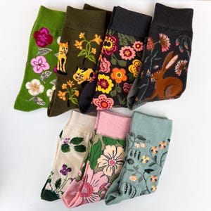 Può includere: Una collezione di calzini colorati con vari motivi. I disegni includono composizioni floreali, gatti e un coniglio. I calzini sono nelle tonalità di verde, nero, rosa e beige. I calzini sono in materiale morbido.