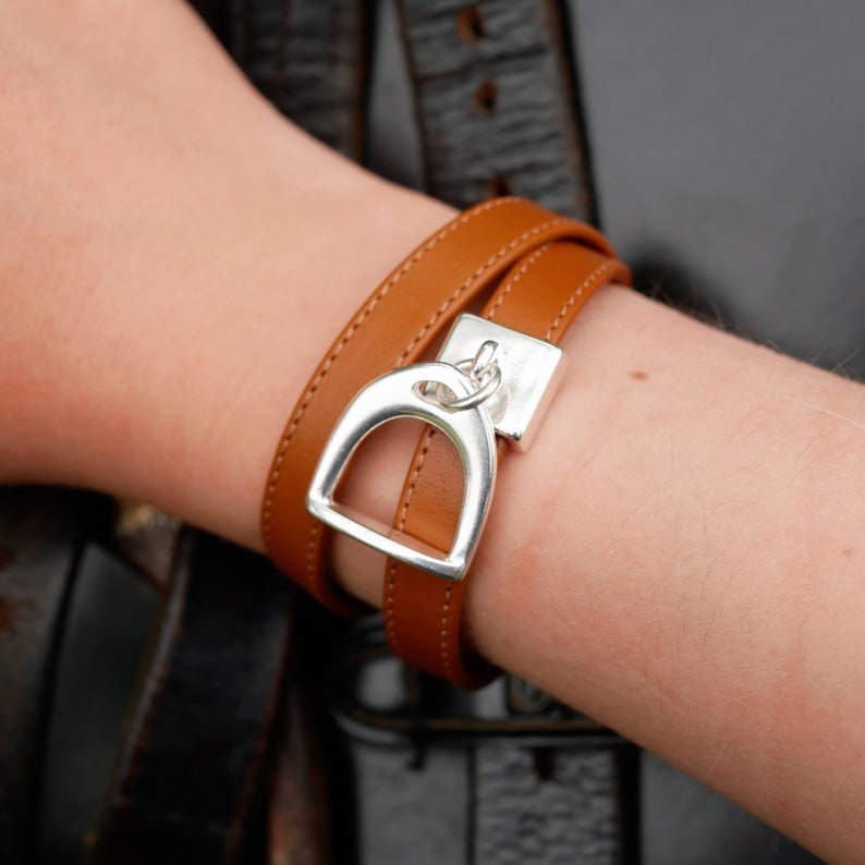 Pode incluir: Um bracelete de couro marrom de dupla volta com um fecho de estribo prateado. O bracelete &eacute; mostrado no pulso de uma pessoa.