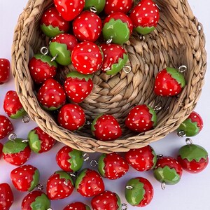 Peut inclure: Des breloques en forme de fraises rouges à pois blancs et chapeaux verts, certaines dans un panier en osier. Les breloques ont des boucles argentées pour les attacher à des bijoux ou autres créations. Les fraises sont éparpillées sur une surface blanche.
