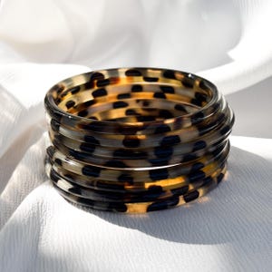 Op de afbeelding: Stapel van zes doorschijnende armbanden met een schildpadpatroon. De armbanden zijn bruin, zwart en beige en zijn op elkaar gestapeld. De achtergrond is een witte, getextureerde stof.