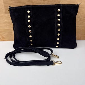 May include: Black suede clutch with gold-colored stud accents and a detachable shoulder strap. The rectangular bag features vertical rows of studs and gold-toned hardware on the strap.