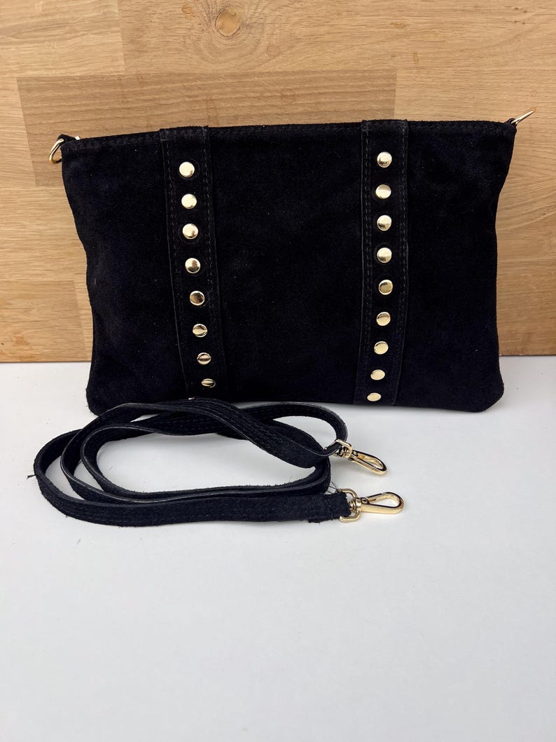 May include: Black suede clutch with gold-colored stud accents and a detachable shoulder strap. The rectangular bag features vertical rows of studs and gold-toned hardware on the strap.