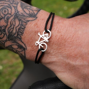 Peut inclure: Un bracelet en cordon noir avec un pendentif en argent en forme de vélo. Le bracelet est porté au poignet.