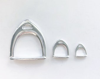Pode incluir: Três pingentes de metal prateado em forma de estribo de diferentes tamanhos. O maior pingente mede aproximadamente 5 cm de altura e o menor mede aproximadamente 2,5 cm de altura.