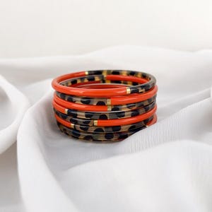 Può includere: Pila di sei braccialetti. I braccialetti alternano tra un arancione brillante e un motivo maculato. I braccialetti con stampa leopardata hanno macchie nere su sfondo trasparente. Sono visibili dettagli dorati. I braccialetti sono su una superficie bianca.