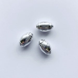 May include: Three silver football-shaped beads. The beads are smooth and have a raised design that resembles the laces of a football.