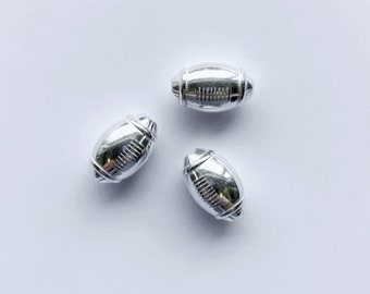 May include: Three silver football-shaped beads. The beads are smooth and have a raised design that resembles the laces of a football.