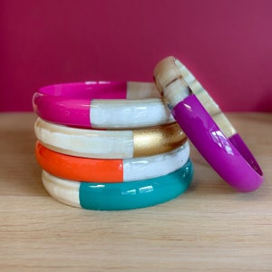 May include: Stack of five colorful bangles with a white base and a different color on the top half of each bangle. The colors are pink, gold, orange, and teal.