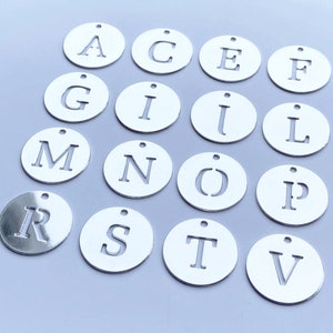 Op de afbeelding: Een set zilveren metalen letterbedels met een gat om op te hangen. De letters zijn A, C, E, F, G, I, J, L, M, N, O, P, R, S, T en V.