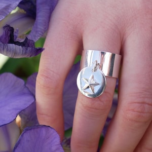May include: A silver ring with a star-shaped charm hanging from it. The ring is wide and has a smooth, polished finish. The star charm is also silver and has a three-dimensional design.