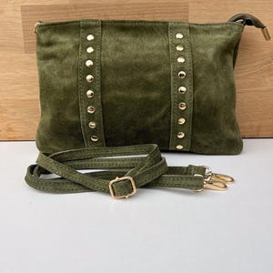 May include: Olive green suede crossbody bag with gold-colored stud accents and hardware. The bag features a detachable shoulder strap with a gold buckle and clasps. The bag is rectangular in shape.
