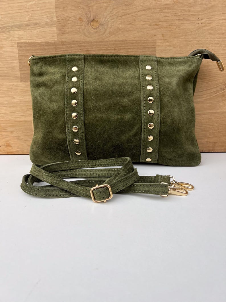 May include: Olive green suede crossbody bag with gold-colored stud accents and hardware. The bag features a detachable shoulder strap with a gold buckle and clasps. The bag is rectangular in shape.