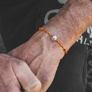Könnte beinhalten: Ein Armband aus orangefarbenem Kordel mit einem runden Silberanhänger. Der Anhänger ist eine runde Kugel mit einer strukturierten Oberfläche.
