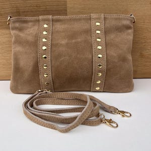 May include: A tan suede clutch with gold-colored studs and hardware. The rectangular bag has a detachable shoulder strap. The bag is displayed on a white surface with a wooden background.