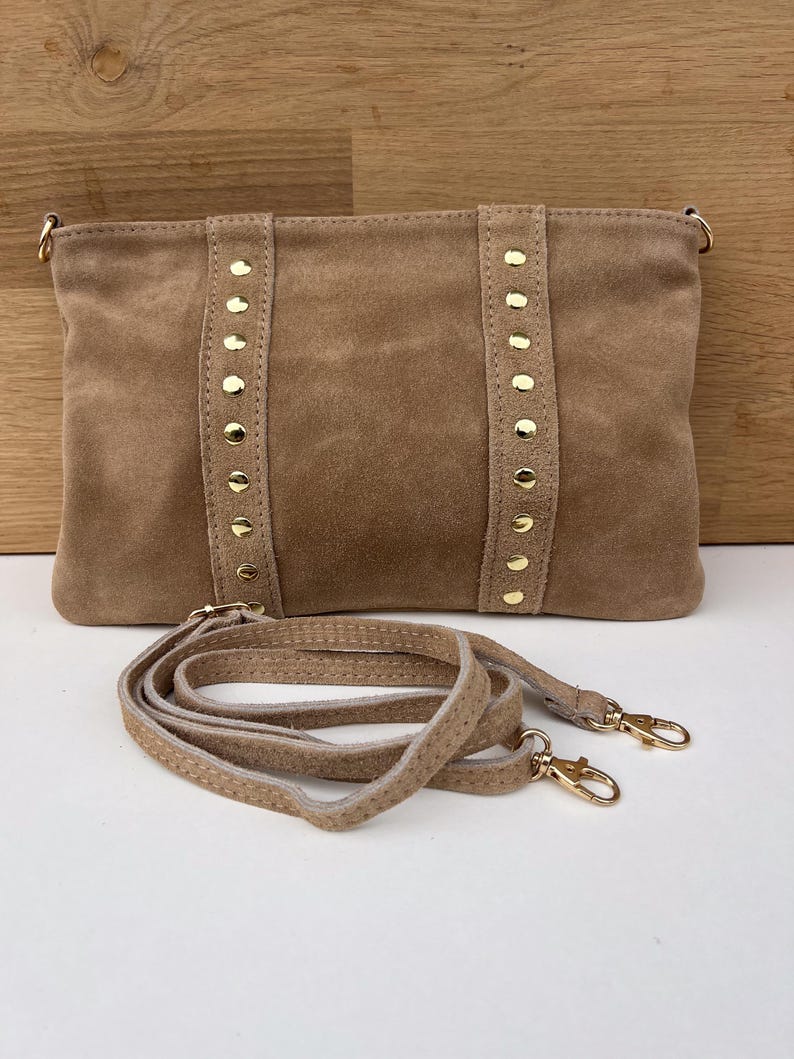 May include: A tan suede clutch with gold-colored studs and hardware. The rectangular bag has a detachable shoulder strap. The bag is displayed on a white surface with a wooden background.