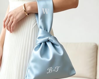 May include: A light blue satin knot bag featuring a wrist handle with the letter W embroidered in white and the text B+T embroidered in white on the main body of the bag. The bag has a smooth, lustrous finish and a soft, draped structure.