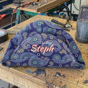 May include: A blue fabric cap with a repeating paisley pattern in shades of yellow, green, and light blue. The word Cycling is embroidered in white above the name Steph, which is stitched in a red script font on the front of the cap.