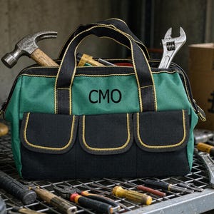 May include: A green and black canvas tool bag featuring three exterior pockets with yellow trim and the letters CMO printed in black on the front. The bag has two black handles and contains a metal hammer and an adjustable wrench inside.