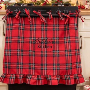 May include: A red and black tartan fabric oven cover featuring a ruffled bottom edge and fabric ties at the top. The text "Natalie's Kitchen" is printed in black script and block lettering in the centre of the fabric, which hangs over a black stove.