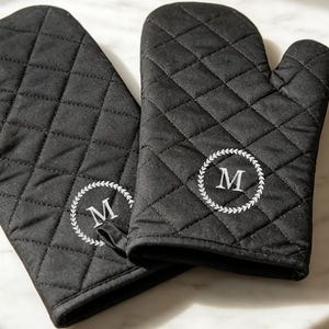 Puede incluir: Un par de manoplas de cocina negras acolchadas con un diseño de corona de laurel circular bordado en blanco y la letra M centrada en cada manopla. La tela presenta un patrón de costuras en forma de diamante en toda la superficie exterior.