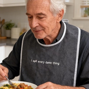 Peut inclure: Une bavette pour adulte gris foncé avec une bordure blanche et le texte "I spill every damn thing" brodé en blanc. La bavette est portée sur une chemise à manches longues gris foncé, couvrant la poitrine pendant le repas dans une assiette.