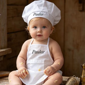 May include: A white chef hat and a matching white apron, both embroidered with the name Paular in black script. The apron features a front pocket and is worn by a baby sitting on a wooden table covered in flour, cookie shapes, and butter.